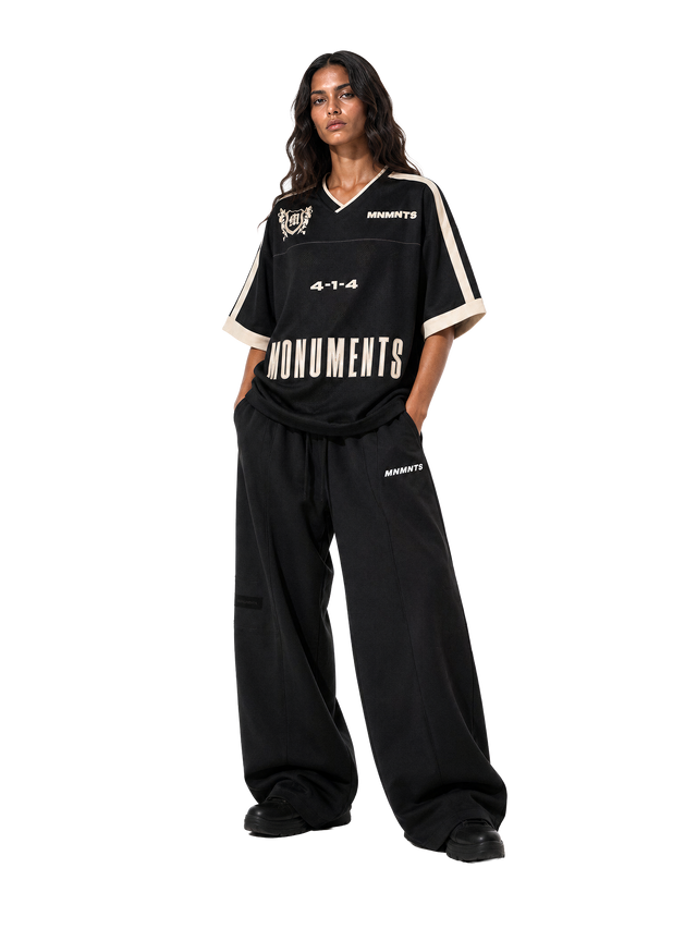 Model wearing the MONUMENTS 4-1-4 Boxy Oversized Jersey in black mesh with eggshell trim and black wide-leg sweatpants, front view.